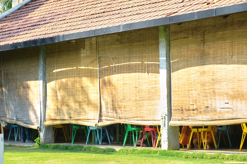 An outdoor seating area with bamboo blinds partially rolled down, revealing colorful chairs underneath. The structure has a tiled roof and is supported by vertical stone pillars. Sunlight casts shadows on the bamboo blinds, creating a warm and inviting ambiance.
