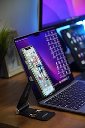 A smartphone is mounted on a black adjustable stand on a wooden desk, displaying a variety of apps on its screen. Behind it is a laptop with a keyboard visible and a tablet with a screen full of icons, all positioned in a modern workspace setup. A green plant is partially visible to the side, adding a touch of natural decor.
