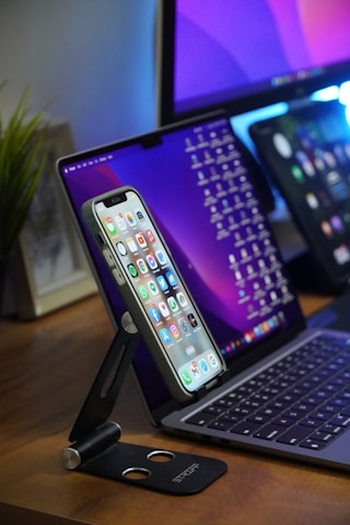 A smartphone is mounted on a black adjustable stand on a wooden desk, displaying a variety of apps on its screen. Behind it is a laptop with a keyboard visible and a tablet with a screen full of icons, all positioned in a modern workspace setup. A green plant is partially visible to the side, adding a touch of natural decor.