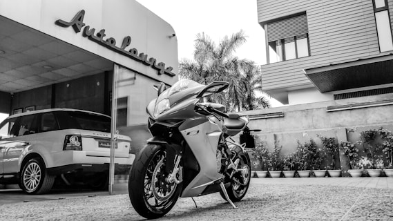 A sleek motorcycle is parked in front of a building with a sign that reads 'Auto Lounge'. To the left, a luxury SUV is visible within a modern showroom. The exterior features contemporary architectural design, complete with large windows and decorative plants lining the entrance. The scene blends elements of high-end automotive culture and modern architecture.