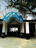 The entrance to a building with a blue semicircular sign written in Hindi. Two black motorcycles are parked in front of the entrance, which is supported by white columns. Trees with sparse foliage arch above the sign, creating a shaded area.