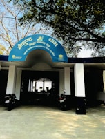 The entrance to a building with a blue semicircular sign written in Hindi. Two black motorcycles are parked in front of the entrance, which is supported by white columns. Trees with sparse foliage arch above the sign, creating a shaded area.
