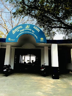The entrance to a building with a blue semicircular sign written in Hindi. Two black motorcycles are parked in front of the entrance, which is supported by white columns. Trees with sparse foliage arch above the sign, creating a shaded area.