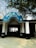 The entrance to a building with a blue semicircular sign written in Hindi. Two black motorcycles are parked in front of the entrance, which is supported by white columns. Trees with sparse foliage arch above the sign, creating a shaded area.