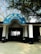 The entrance to a building with a blue semicircular sign written in Hindi. Two black motorcycles are parked in front of the entrance, which is supported by white columns. Trees with sparse foliage arch above the sign, creating a shaded area.