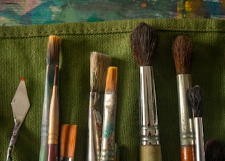 Various paintbrushes and a palette knife are laid on a green canvas surface. The brushes vary in size, shape, and bristle type, indicating use in different painting techniques. The background features a textured green material.