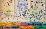 A colorful array of watercolor splatters covers a light surface, with splashes of various hues such as blue, green, yellow, and red scattered throughout. The bottom part of the image features an assortment of watercolor pans with vibrant pigments, arranged in a tray, showcasing different shades.
