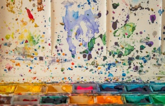 A colorful array of watercolor splatters covers a light surface, with splashes of various hues such as blue, green, yellow, and red scattered throughout. The bottom part of the image features an assortment of watercolor pans with vibrant pigments, arranged in a tray, showcasing different shades.