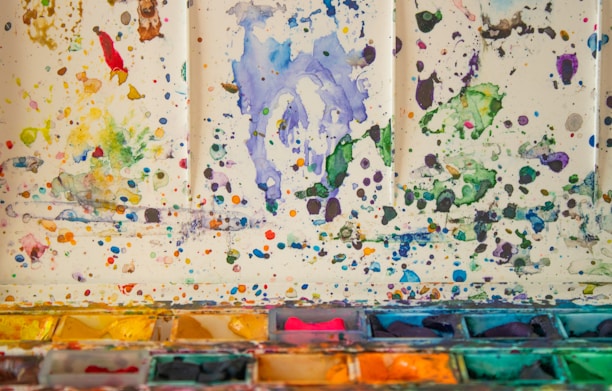 A colorful array of watercolor splatters covers a light surface, with splashes of various hues such as blue, green, yellow, and red scattered throughout. The bottom part of the image features an assortment of watercolor pans with vibrant pigments, arranged in a tray, showcasing different shades.