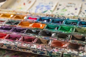 A well-used watercolor palette featuring a variety of vibrant colors, including shades of yellow, red, green, blue, and purple. The paints have been mixed and splattered across the palette, indicating frequent use. The surface of the palette is marked with paint splashes and stains, adding a textured and lively appearance.