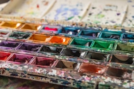 A well-used watercolor palette featuring a variety of vibrant colors, including shades of yellow, red, green, blue, and purple. The paints have been mixed and splattered across the palette, indicating frequent use. The surface of the palette is marked with paint splashes and stains, adding a textured and lively appearance.