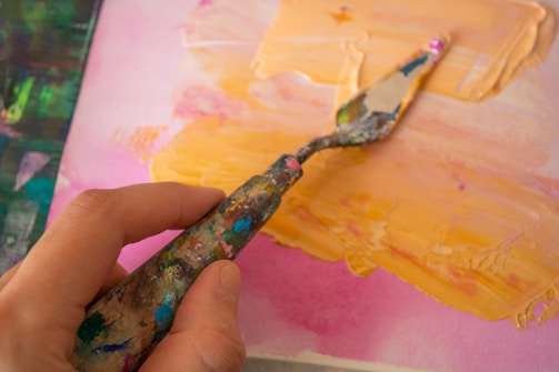 Close-up of an artist’s hand mixing vibrant paints on a palette with energetic brush strokes in the background.