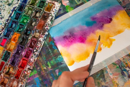 A watercolor palette with a variety of vibrant colors is placed next to a piece of paper featuring a colorful abstract painting. A hand is holding a paintbrush, which is being used to apply paint to the paper. The background is a multicolored surface with remnants of paint, suggesting a creative workspace.