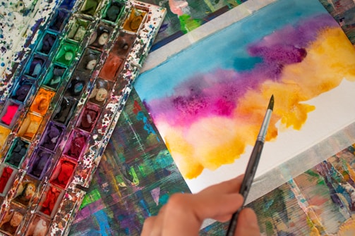 A watercolor palette with a variety of vibrant colors is placed next to a piece of paper featuring a colorful abstract painting. A hand is holding a paintbrush, which is being used to apply paint to the paper. The background is a multicolored surface with remnants of paint, suggesting a creative workspace.