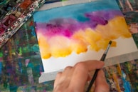 A person is creating an artwork using watercolor paints. The painting features a gradient of colors transitioning from blue and purple at the top to yellow and orange at the bottom. The hand holding the paintbrush is seen in the foreground, while a palette of watercolors is partially visible on the left side of the image.