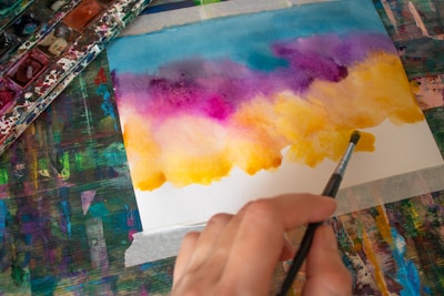 A person is creating an artwork using watercolor paints. The painting features a gradient of colors transitioning from blue and purple at the top to yellow and orange at the bottom. The hand holding the paintbrush is seen in the foreground, while a palette of watercolors is partially visible on the left side of the image.