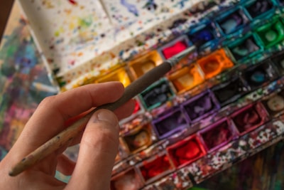 A hand holding a paintbrush hovers over a colorful watercolor palette filled with an array of vibrant hues. The palette is splattered with various colors, indicating frequent use. The scene captures the tools of an artist in mid-creation.