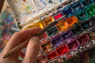 A hand holding a paintbrush hovers over a colorful watercolor palette filled with an array of vibrant hues. The palette is splattered with various colors, indicating frequent use. The scene captures the tools of an artist in mid-creation.