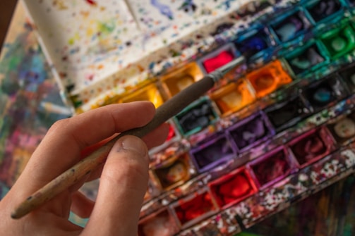 A hand holding a paintbrush hovers over a colorful watercolor palette filled with an array of vibrant hues. The palette is splattered with various colors, indicating frequent use. The scene captures the tools of an artist in mid-creation.