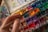 A hand holding a paintbrush hovers over a colorful watercolor palette filled with an array of vibrant hues. The palette is splattered with various colors, indicating frequent use. The scene captures the tools of an artist in mid-creation.