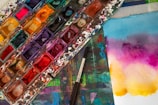 A vibrant and colorful watercolor palette sits on a stained surface, accompanied by a brush. Nearby, a piece of paper displays a gradient of bright colors blending smoothly from blue to purple to yellow.