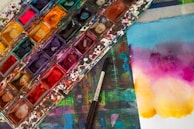 A vibrant and colorful watercolor palette sits on a stained surface, accompanied by a brush. Nearby, a piece of paper displays a gradient of bright colors blending smoothly from blue to purple to yellow.