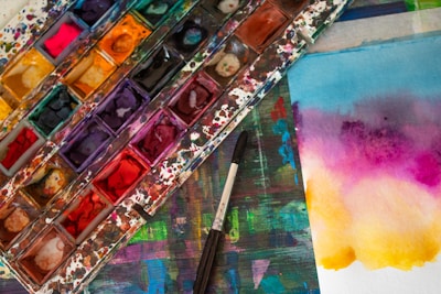 A vibrant and colorful watercolor palette sits on a stained surface, accompanied by a brush. Nearby, a piece of paper displays a gradient of bright colors blending smoothly from blue to purple to yellow.
