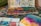 An artist's workspace features a well-used watercolor palette with a variety of vibrant colors. The table is covered in paint splatters and artistic tools, including paintbrushes. A watercolor painting in progress displays a blend of rich pink, yellow, and blue hues.