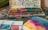 An artist's workspace features a well-used watercolor palette with a variety of vibrant colors. The table is covered in paint splatters and artistic tools, including paintbrushes. A watercolor painting in progress displays a blend of rich pink, yellow, and blue hues.