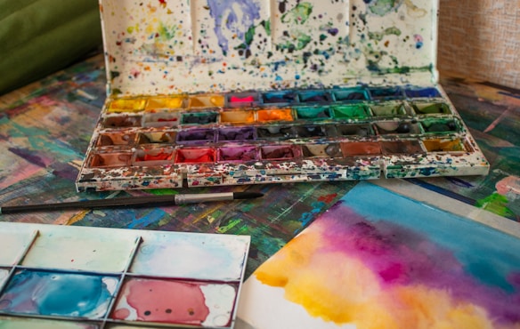 An artist's workspace features a well-used watercolor palette with a variety of vibrant colors. The table is covered in paint splatters and artistic tools, including paintbrushes. A watercolor painting in progress displays a blend of rich pink, yellow, and blue hues.