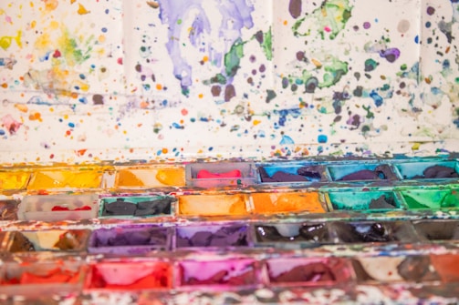A vibrant assortment of watercolor paints in a palette, with splatters and stains covering the surrounding area. The array consists of various shades including yellows, oranges, reds, greens, blues, and purples. The background is filled with colorful splatters creating a chaotic yet artistic effect.