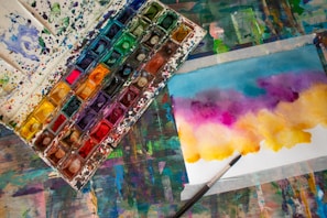 A watercolor paint palette filled with various vibrant colors is placed next to a piece of paper showcasing an abstract artwork. The artwork features a blend of blue, purple, pink, and yellow hues. A paintbrush lies beside the paper, indicating an ongoing painting process. The entire scene is set on a colorful, paint-splattered table, adding an artistic atmosphere.