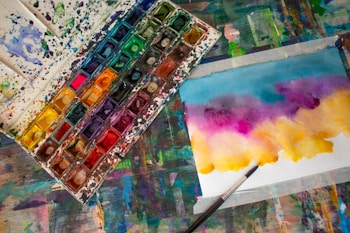 A watercolor paint palette filled with various vibrant colors is placed next to a piece of paper showcasing an abstract artwork. The artwork features a blend of blue, purple, pink, and yellow hues. A paintbrush lies beside the paper, indicating an ongoing painting process. The entire scene is set on a colorful, paint-splattered table, adding an artistic atmosphere.