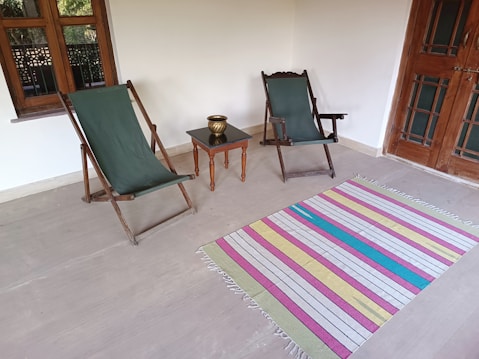 Two green canvas deck chairs and a small wooden table with a brass pot are arranged on a minimalist porch. A colorful striped rug lies on the floor between them. The space includes wooden windows and a door, giving a rustic and cozy feel.