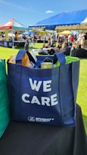 A blue tote bag with the words 'WE CARE' and 'RIVERCITY CHURCH' printed on it, filled with various items including snacks. The bag is placed on a black surface in an outdoor event setting. In the background, there are open tents with people sitting on the grass, enjoying the sunny weather.