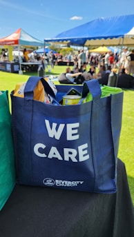 A blue tote bag with the words 'WE CARE' and 'RIVERCITY CHURCH' printed on it, filled with various items including snacks. The bag is placed on a black surface in an outdoor event setting. In the background, there are open tents with people sitting on the grass, enjoying the sunny weather.