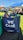 A blue tote bag with the words 'WE CARE' and 'RIVERCITY CHURCH' printed on it, filled with various items including snacks. The bag is placed on a black surface in an outdoor event setting. In the background, there are open tents with people sitting on the grass, enjoying the sunny weather.