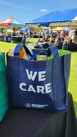 A blue tote bag with the words 'WE CARE' and 'RIVERCITY CHURCH' printed on it, filled with various items including snacks. The bag is placed on a black surface in an outdoor event setting. In the background, there are open tents with people sitting on the grass, enjoying the sunny weather.
