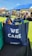 A blue tote bag with the words 'WE CARE' and 'RIVERCITY CHURCH' printed on it, filled with various items including snacks. The bag is placed on a black surface in an outdoor event setting. In the background, there are open tents with people sitting on the grass, enjoying the sunny weather.