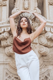 A person stands confidently against an ornate stone backdrop, with arms raised above their head. The person is dressed in a brown sleeveless top and white pants, exuding a sense of elegance and poise.