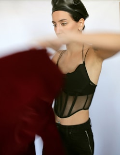 A person wearing a black mesh top and a leather beret appears to be in motion, holding a red garment. The setting is minimalistic with a blurred background.