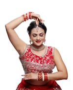 A person dressed in traditional attire is performing a dance pose. The outfit is richly decorated with intricate designs and vibrant red color. Jewelry adorns the individual, including earrings, a necklace, a headpiece, and multiple bangles. The expression is serene, with closed eyes and a slight smile.