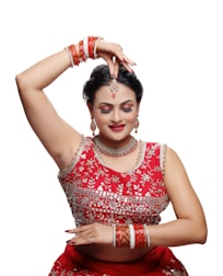 A person dressed in traditional attire is performing a dance pose. The outfit is richly decorated with intricate designs and vibrant red color. Jewelry adorns the individual, including earrings, a necklace, a headpiece, and multiple bangles. The expression is serene, with closed eyes and a slight smile.