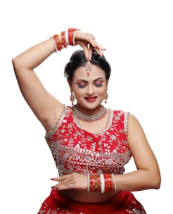 A person dressed in traditional attire is performing a dance pose. The outfit is richly decorated with intricate designs and vibrant red color. Jewelry adorns the individual, including earrings, a necklace, a headpiece, and multiple bangles. The expression is serene, with closed eyes and a slight smile.