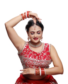 A person dressed in traditional attire is performing a dance pose. The outfit is richly decorated with intricate designs and vibrant red color. Jewelry adorns the individual, including earrings, a necklace, a headpiece, and multiple bangles. The expression is serene, with closed eyes and a slight smile.