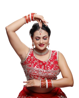 A person dressed in traditional attire is performing a dance pose. The outfit is richly decorated with intricate designs and vibrant red color. Jewelry adorns the individual, including earrings, a necklace, a headpiece, and multiple bangles. The expression is serene, with closed eyes and a slight smile.