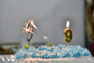 A birthday cake decorated with blue and white frosting features two sparklers, forming the number 9. One is sparking while the other burns steadily. Small decorative pearls are scattered over the icing.