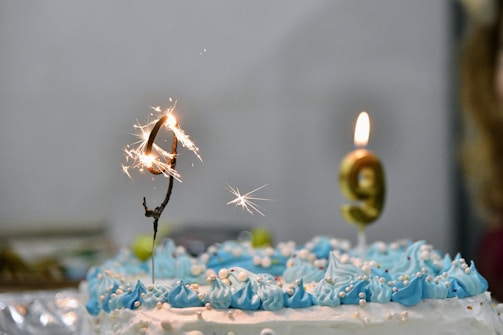 A birthday cake decorated with blue and white frosting features two sparklers, forming the number 9. One is sparking while the other burns steadily. Small decorative pearls are scattered over the icing.