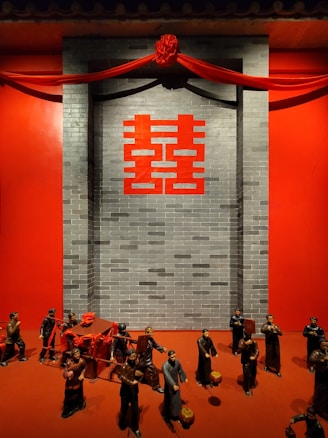 Clay figurines are arranged in front of a gray brick wall with a large red double happiness symbol. The figures seem to be part of a traditional Chinese procession, carrying a red sedan chair and holding various objects, such as lanterns and banners. Above them, red draped fabric and a decorative knot enhance the festive atmosphere.