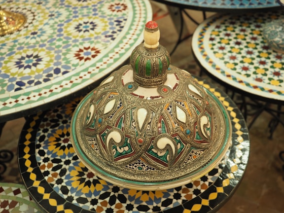An intricately designed decorative Moroccan tagine lid rests on a colorful mosaic table. The lid is adorned with detailed geometric patterns and an array of colors, including green, blue, and red accents against a metallic and ivory background. Surrounding the tagine are several other tables covered with similarly vibrant mosaic patterns.